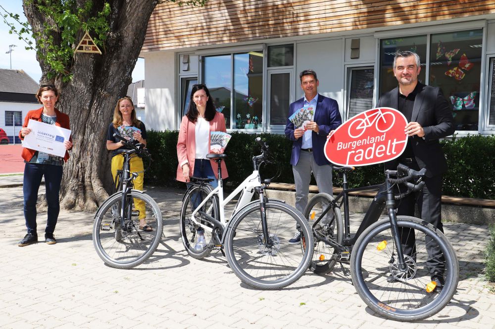 5 Personen mit Fahrrädern und dem Burgenland radelt Logo als Tafel vor einer Schule