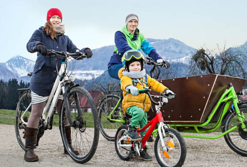 Pärchen mit Kleinkind fahren mit Lastenrad und Rad vor verschneiten Bergen